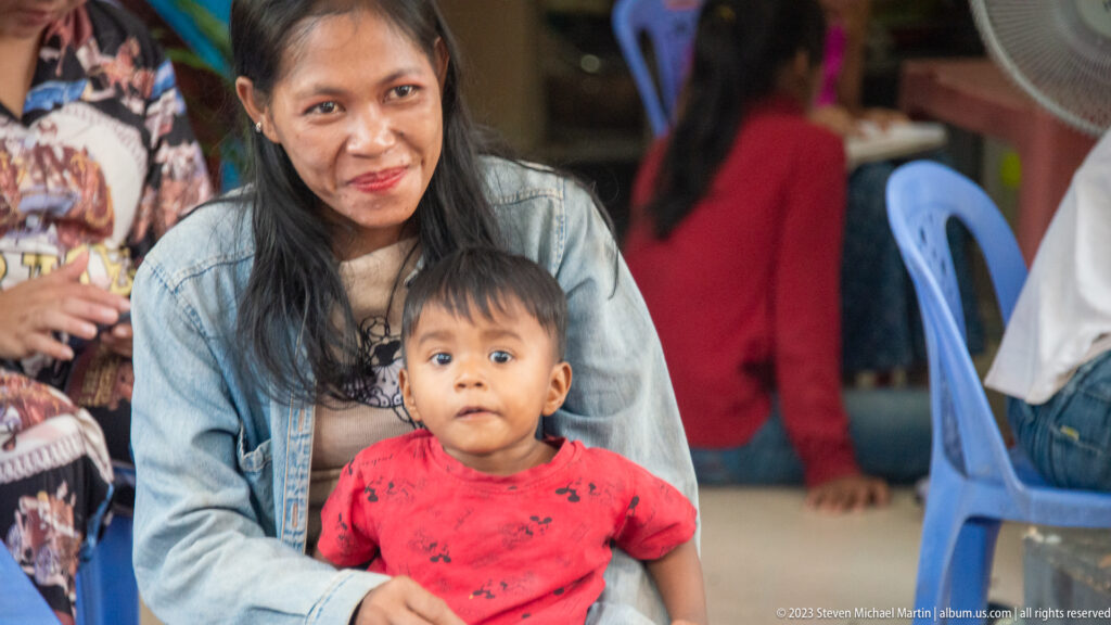 Seated woman holding a young child on her lap, both looking toward the camera in a casual local setting.