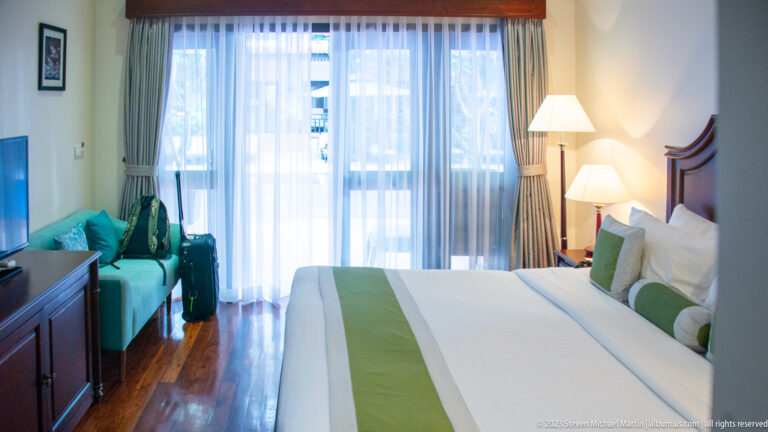 Hotel room viewed toward sheer balcony curtains, with bed at right, turquoise bench, luggage, and wooden cabinet at left.