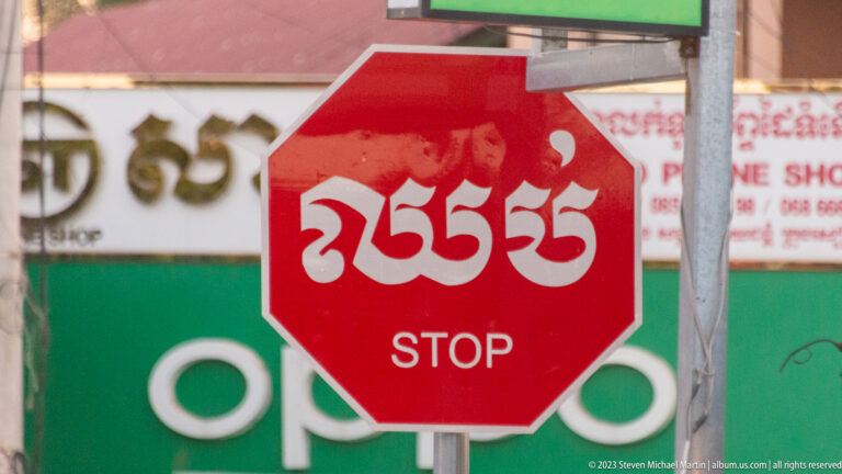 Red octagonal stop sign with white Khmer script above the English word STOP, set against storefront signs.