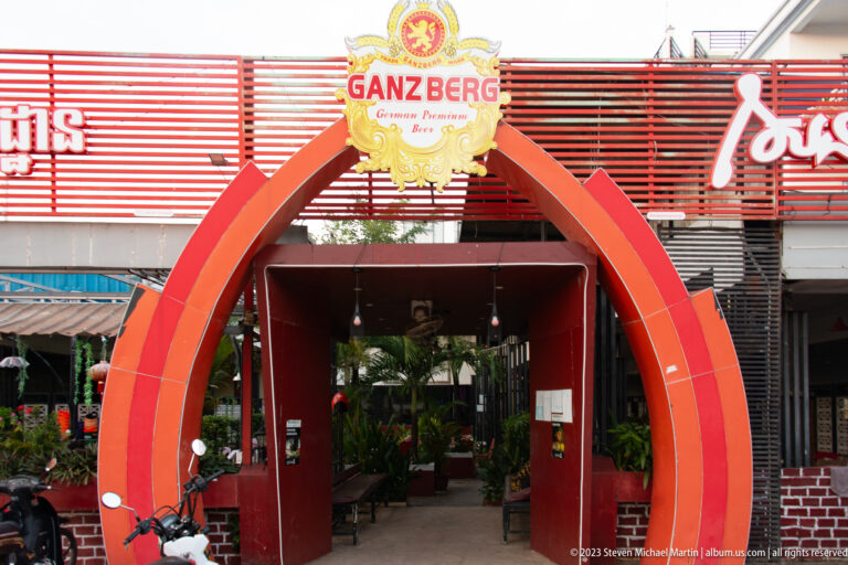 Red arched restaurant entrance with Ganzberg beer sign, Khmer lettering, and parked motorbikes along the street.