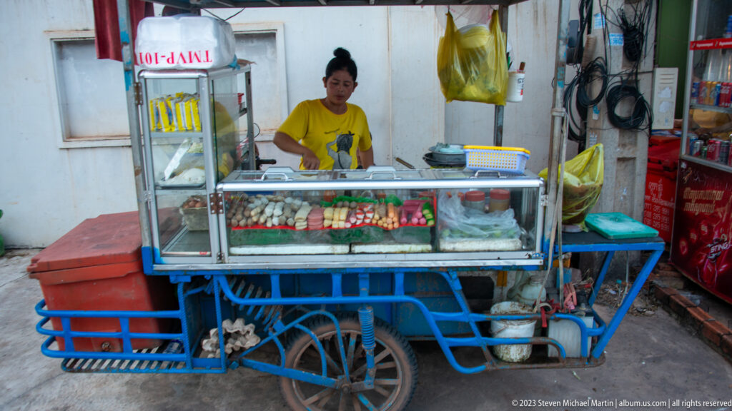 Woman standing behind a blue street-food cart with glass display, eggs, cooking equipment, and hanging supplies.
