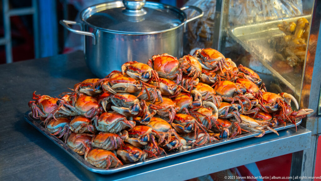 Pile of orange-red cooked crabs on a metal tray beside a large cooking pot at a street-food stall.