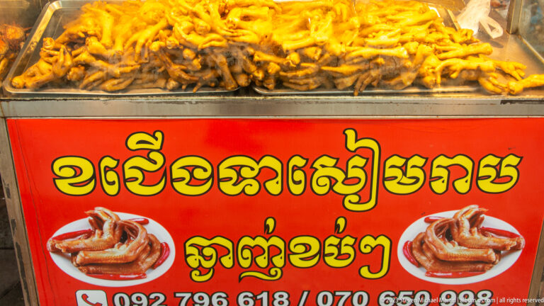 Red sign with large yellow Khmer lettering, fried-food photographs, phone numbers, and trays of food above.