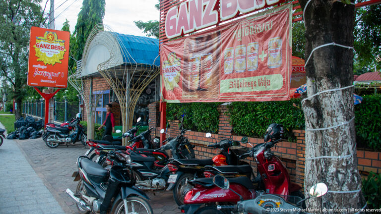 Rows of parked motorbikes beneath red Ganzberg beer signs outside a roadside venue with trees and brick walls.