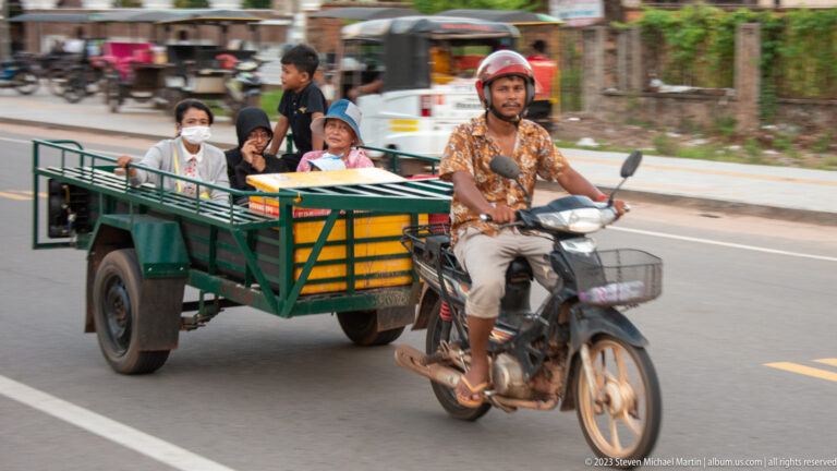 Motorcyclist pulling an open green trailer with several seated passengers along a paved road in Siem Reap.