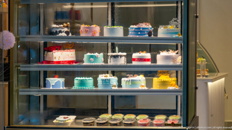 Refrigerated bakery case filled with colorful frosted cakes, tiered designs, and small individual desserts.