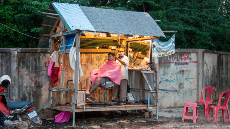 Customer receiving a haircut in a small wooden roadside barbershop with corrugated roof and plastic chairs.