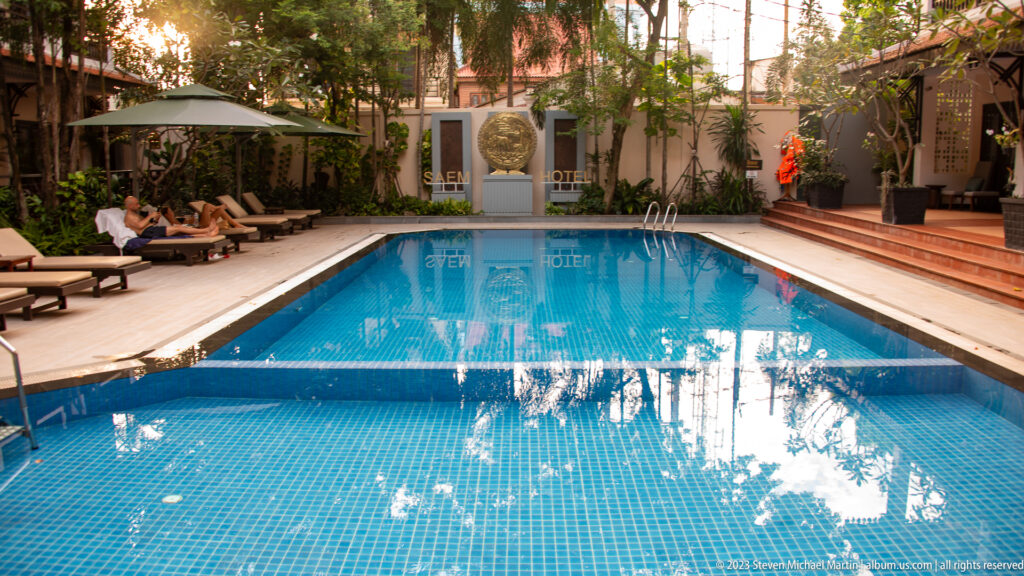 Rectangular blue hotel pool bordered by wooden decking, lounge chairs, trees, and shaded courtyard seating.
