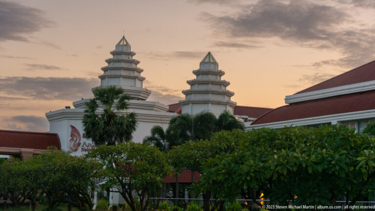Twin stepped hotel towers and red roofs rising above palm trees beneath a cloudy dusk sky.
