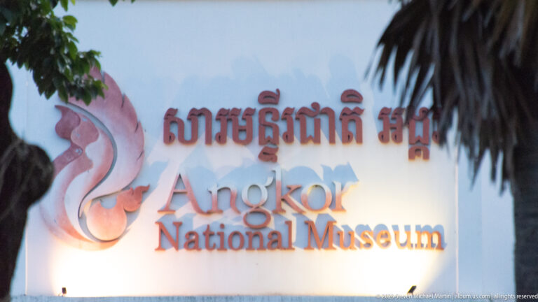 Illuminated Angkor National Museum sign in Khmer and English, partly framed by dark palm fronds.