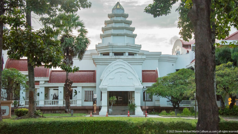 White museum façade with tiered central tower, red tile roofs, arched entrance, lawn, trees, and hedges.