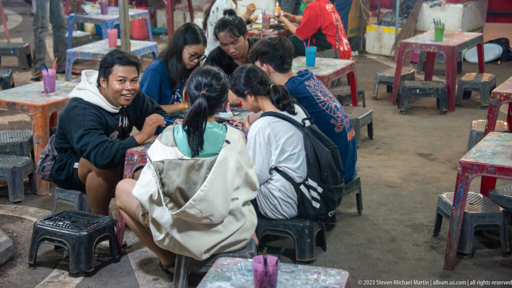 Group of young diners seated on low plastic stools around a table in a busy open-air food venue.