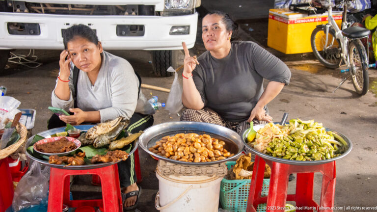 Two seated women behind bowls and trays of prepared foods, vegetables, eggs, and sauces at a market stall.
