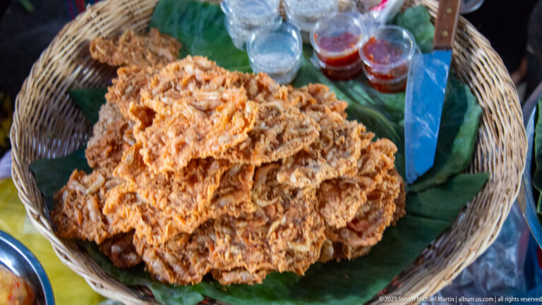 Large mound of crispy shredded fried food on banana leaves in a woven basket, with dipping sauces behind.
