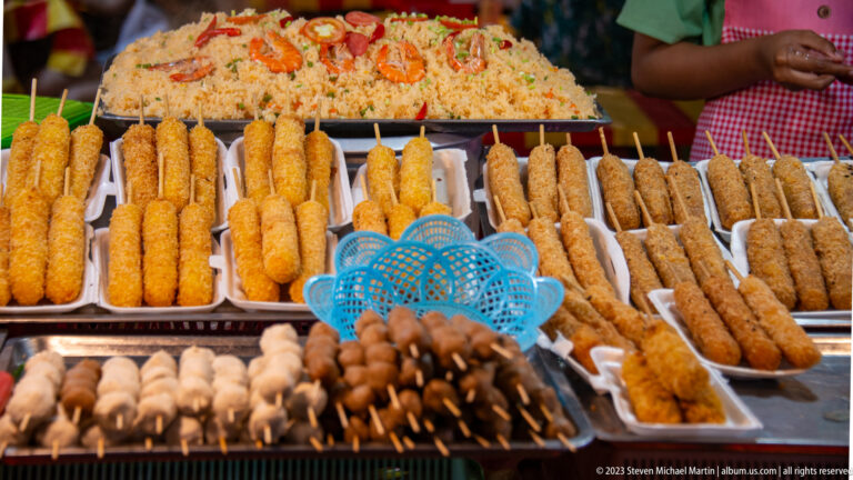 Rows of assorted breaded and grilled foods on wooden skewers arranged in metal trays at a market stall.