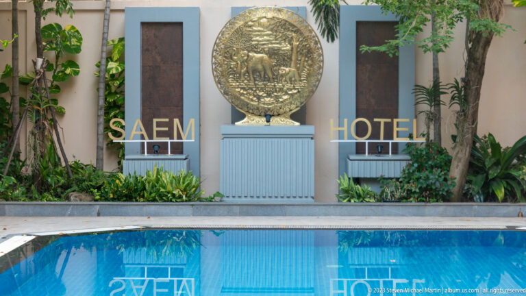 Circular elephant relief mounted between dark wall panels above a hotel pool, with the Saem Hotel name and reflections below.