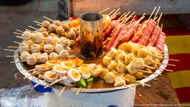 Circular street-food grill with skewered sausages, meatballs, eggs, tofu, and other foods arranged around a central chimney.