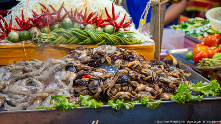 Raw shellfish and seafood piled before tomatoes, cucumbers, limes, chilies, and shredded vegetables at a market stall.