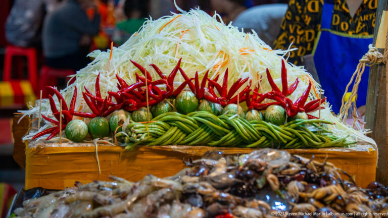 Shredded green papaya stacked above red chilies, green limes, and coiled long beans at a food stall.