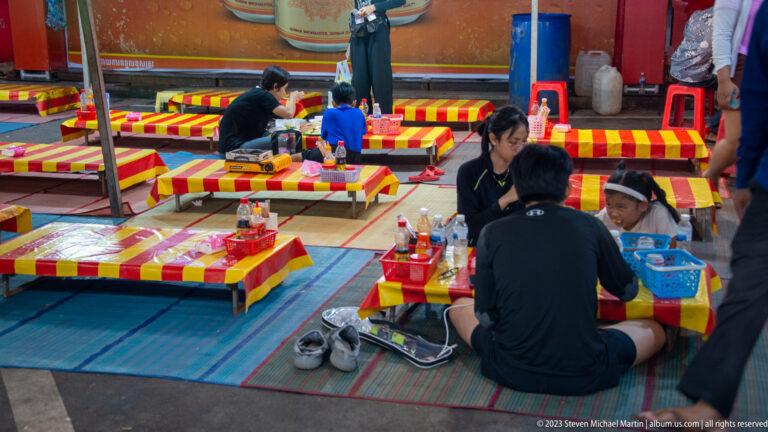 Diners seated on mats around low red-and-yellow tables, with shoes and condiments beside the dining area.