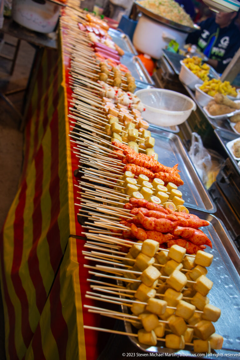 Long diagonal rows of wooden skewers holding sausages, tofu, vegetables, and assorted prepared foods at a market stall.