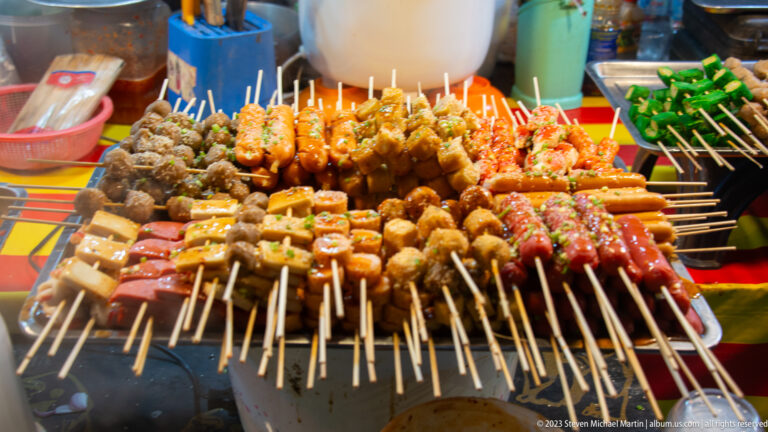 Crowded tray of marinated skewers with meatballs, sausages, tofu, and seasoned meat pieces ready for cooking.