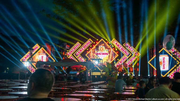 Outdoor night stage with Ganzberg logos, geometric light panels, multicolored beams, and rows of tables.