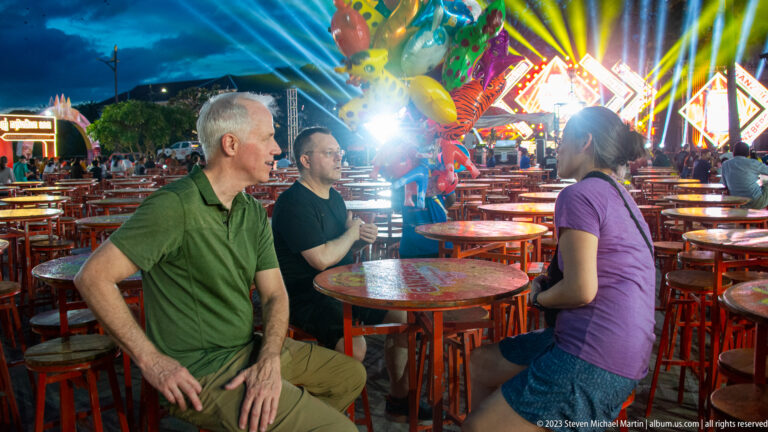 Three adults seated around a round table at a brightly lit outdoor venue with red tables and stage lights.