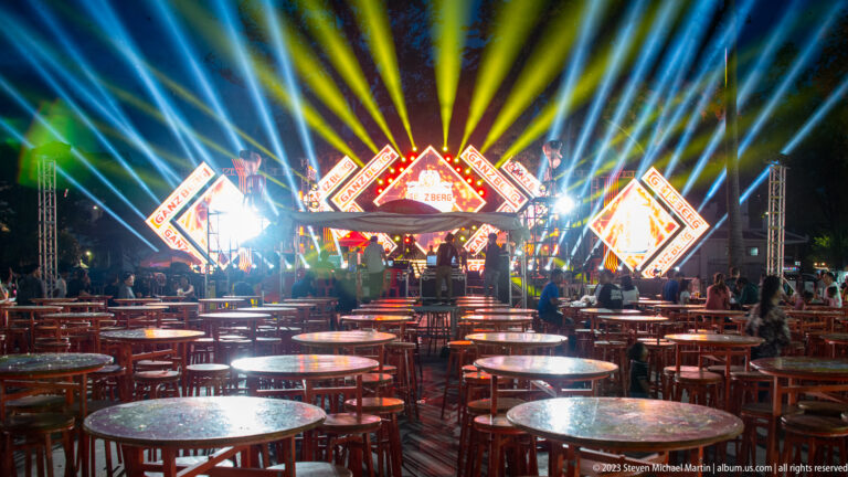 Bright Ganzberg concert stage viewed across rows of round red tables under radiating blue, white, and yellow lights.