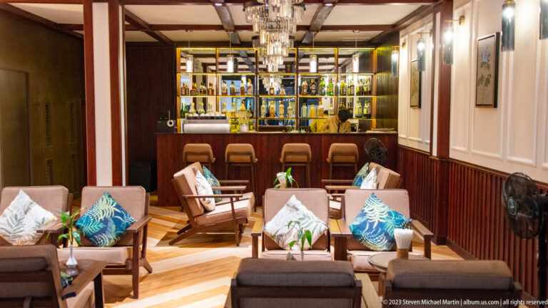 Wood-paneled hotel lounge with sofas, armchairs, tropical cushions, chandelier, and illuminated bar shelves.