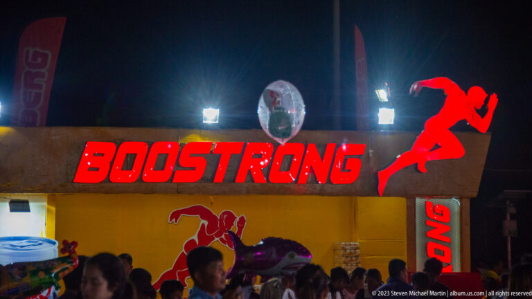 Red illuminated BOOSTRONG sign with running figure logo above a crowded vendor stall at night.