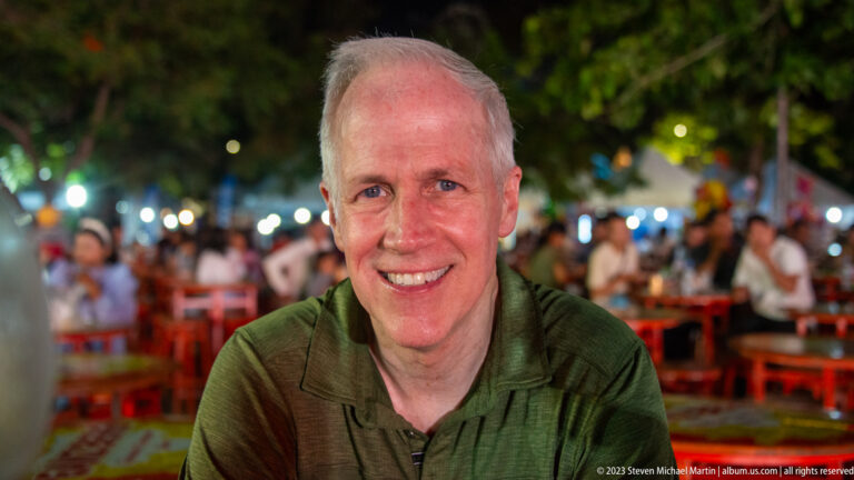 Smiling gray-haired man in a green shirt seated at an outdoor night venue with blurred tables and lights behind.