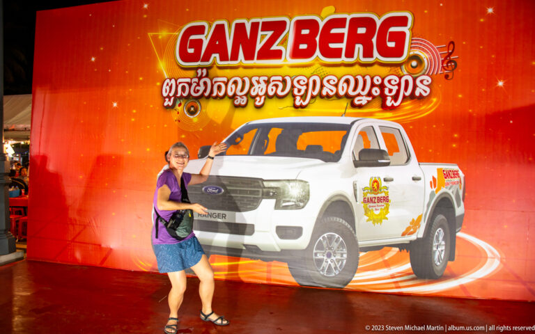 Woman posing beside a red Ganzberg promotional backdrop featuring Khmer text and a white pickup truck.