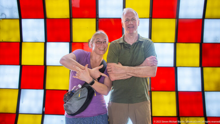 Two smiling adults posing with crossed arms before a grid of illuminated red, yellow, and white panels.