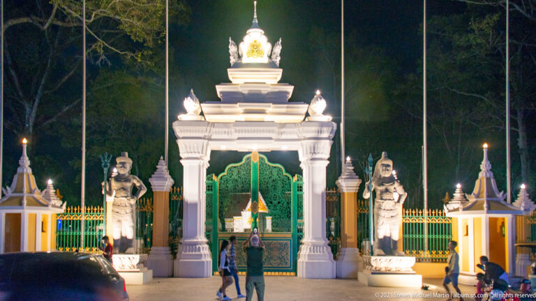 Illuminated white entrance gate with green metal doors, tiered roof forms, and guardian statues on both sides.
