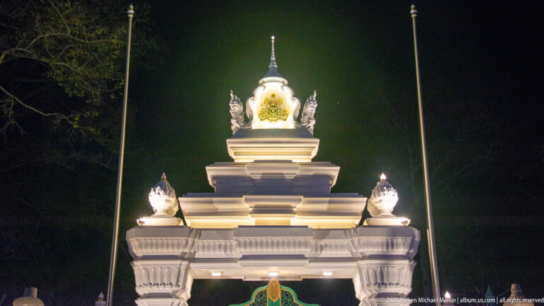 Close view of the tiered upper section of a white illuminated gate with finials and glowing central crest.