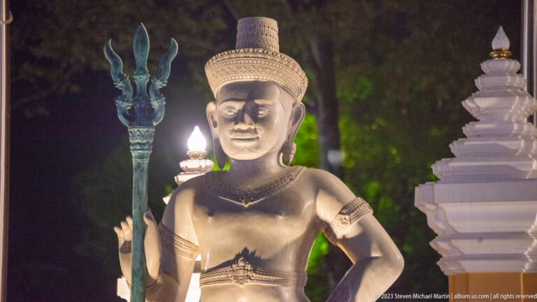 Illuminated stone-colored male guardian statue wearing a tall headdress and holding a vertical staff beside a white gate.