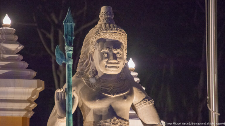 Second illuminated guardian statue with elaborate headdress and vertical staff standing beside the ceremonial gate.