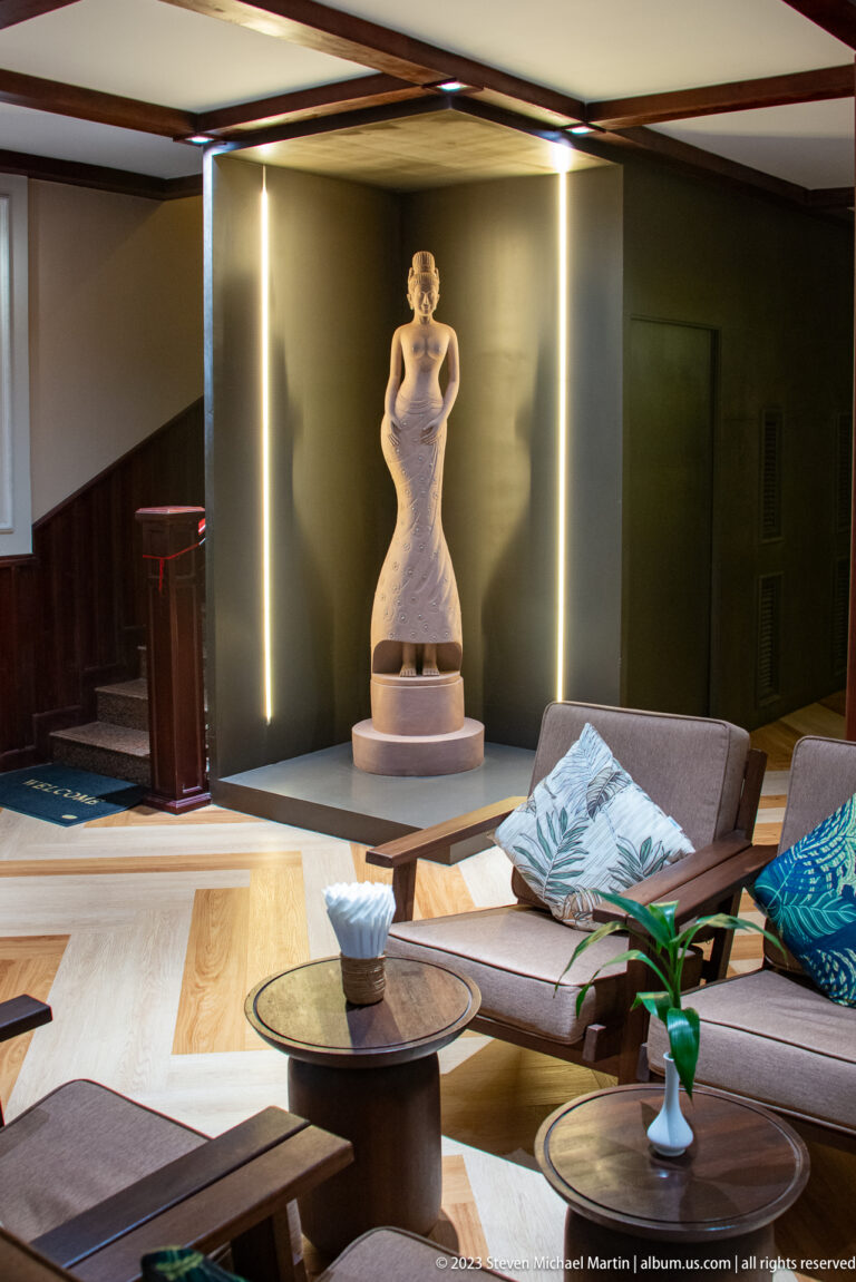 Dim hotel lounge with low seating and tables facing a standing female sculpture illuminated by vertical light strips.