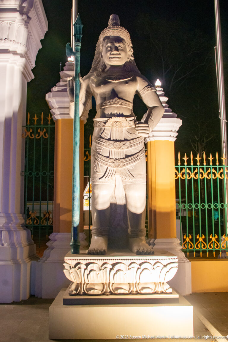 Illuminated guardian statue holding a tall green staff on a lotus pedestal beside an ornate gate.