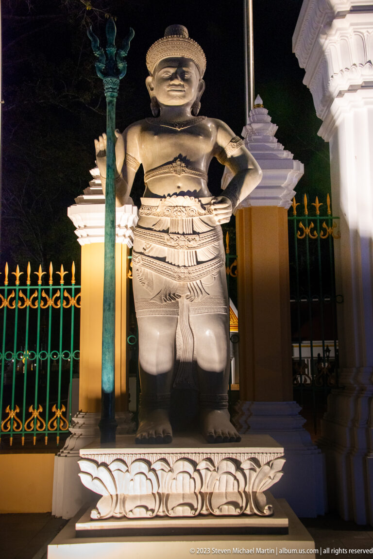 Second illuminated guardian statue with vertical staff, layered costume, and carved pedestal.