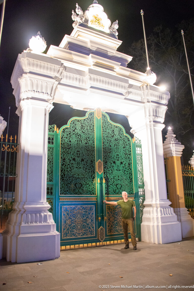 Oblique nighttime view of a white ornamental gateway with green patterned gates and a person at the base.