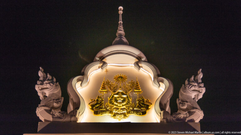 Golden crest set in a white cusped frame with two flanking sculptural creatures at night.