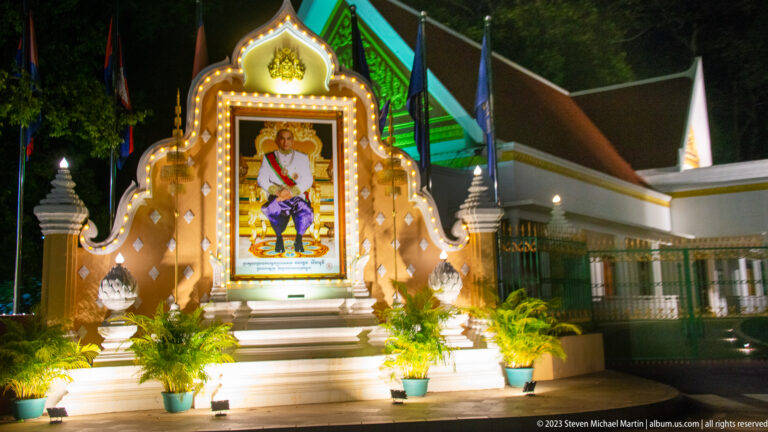 Illuminated portrait of a seated royal figure in an ornate freestanding shrine with steps and potted plants.