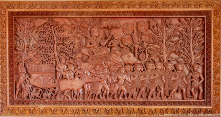 Rectangular carved wooden relief showing a procession, ox-drawn cart, figures, trees, and architectural motifs.