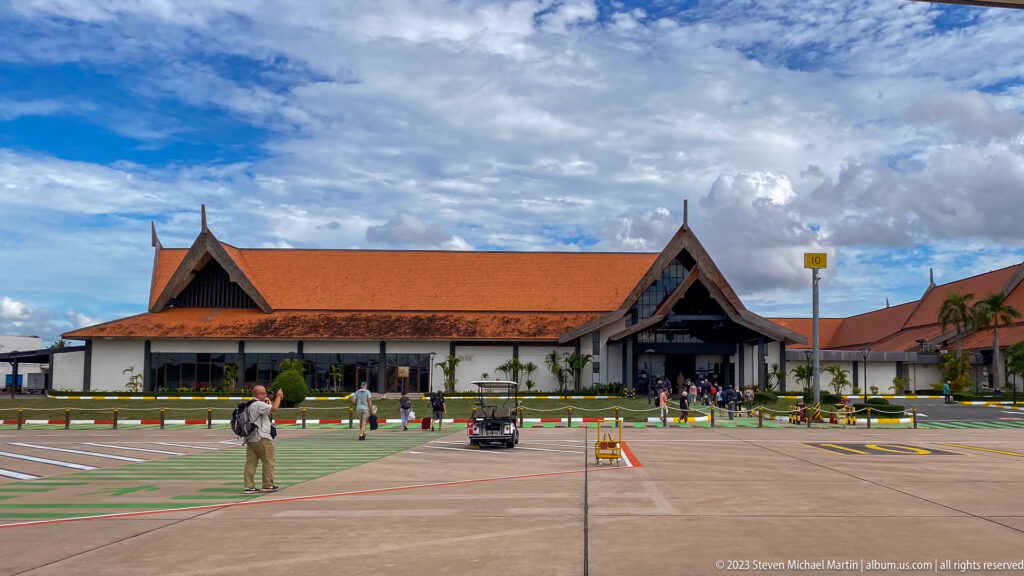 Low airport terminal with red-tiled gabled roofs, passengers, and ground vehicles under a cloudy sky.