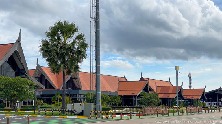 Airport terminal side elevation with steep red roofs, dark gables, palm trees, and service infrastructure.