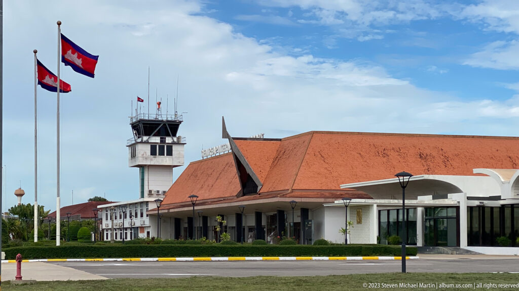 Airport control tower beside a long red-roofed terminal with Cambodian flags and glass-fronted entrance.