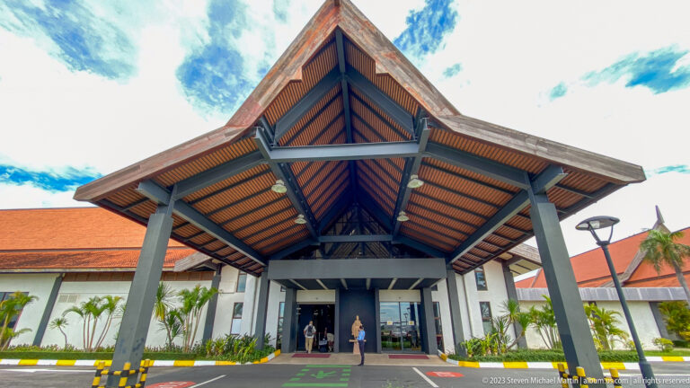 Large peaked airport entrance canopy with dark columns, exposed trusses, and warm-toned roof panels.
