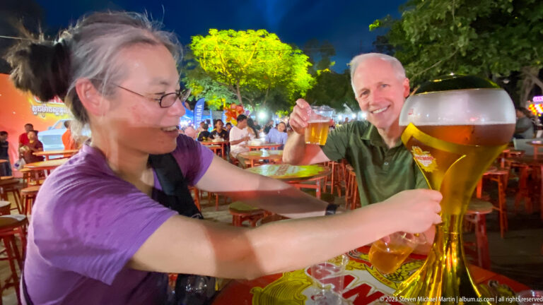 Two smiling visitors gathered around a large beer tower at a brightly lit night market.